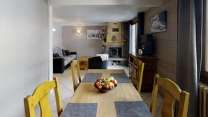 Un coin repas avec une table en bois, des chaises et un panier de fruits. On aperçoit au fond une cheminée, un téléviseur et un canapé.
