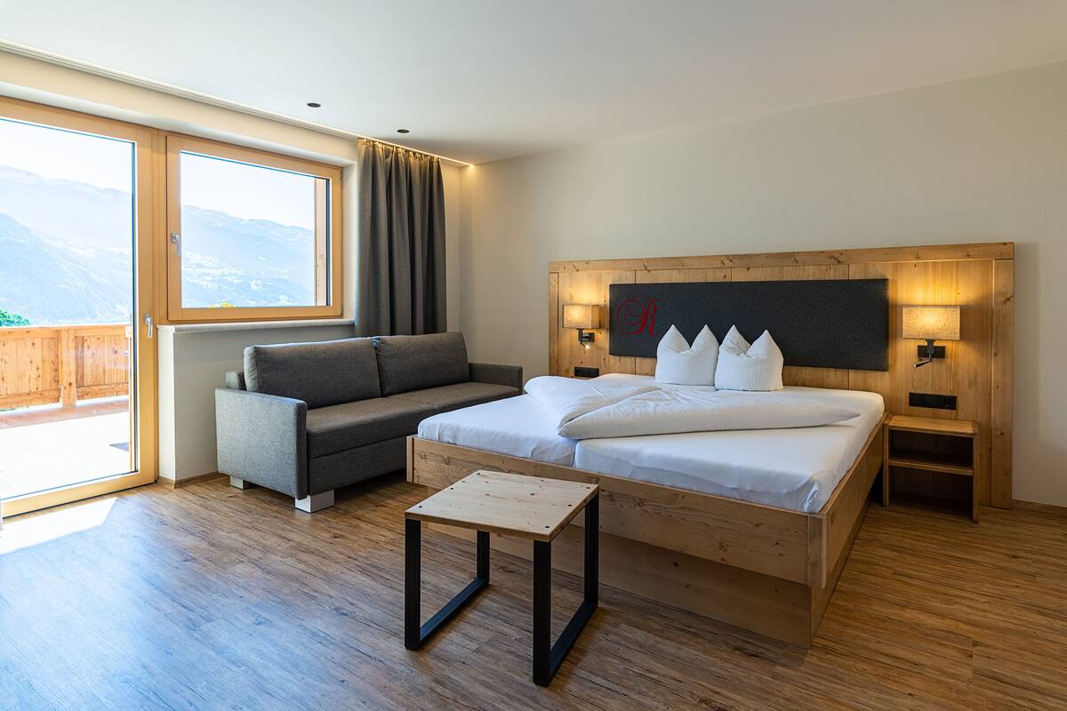 Una habitación de hotel moderna con una cama de madera, un sofá gris, una pequeña mesa de madera y un gran ventanal con vistas a la montaña.