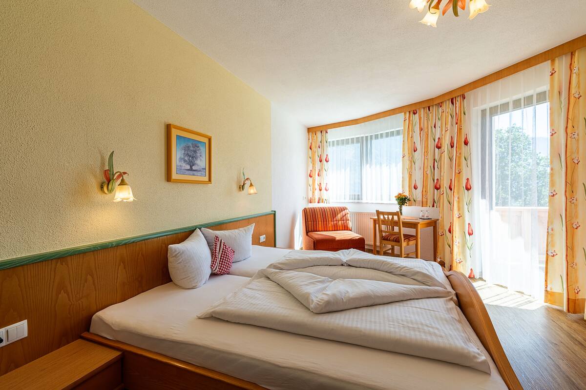Una habitación de hotel con una cama bien tendida, un escritorio pequeño con una silla, una ventana con cortinas y una lámpara fijada a la pared.