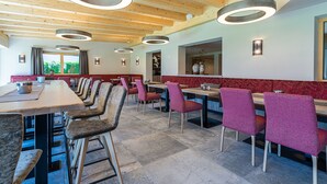 A restaurant with wooden ceiling beams, circular pendant lights, and a mix of red and grey upholstered chairs. - Hotel Garni Romantik (Fuegenberg)