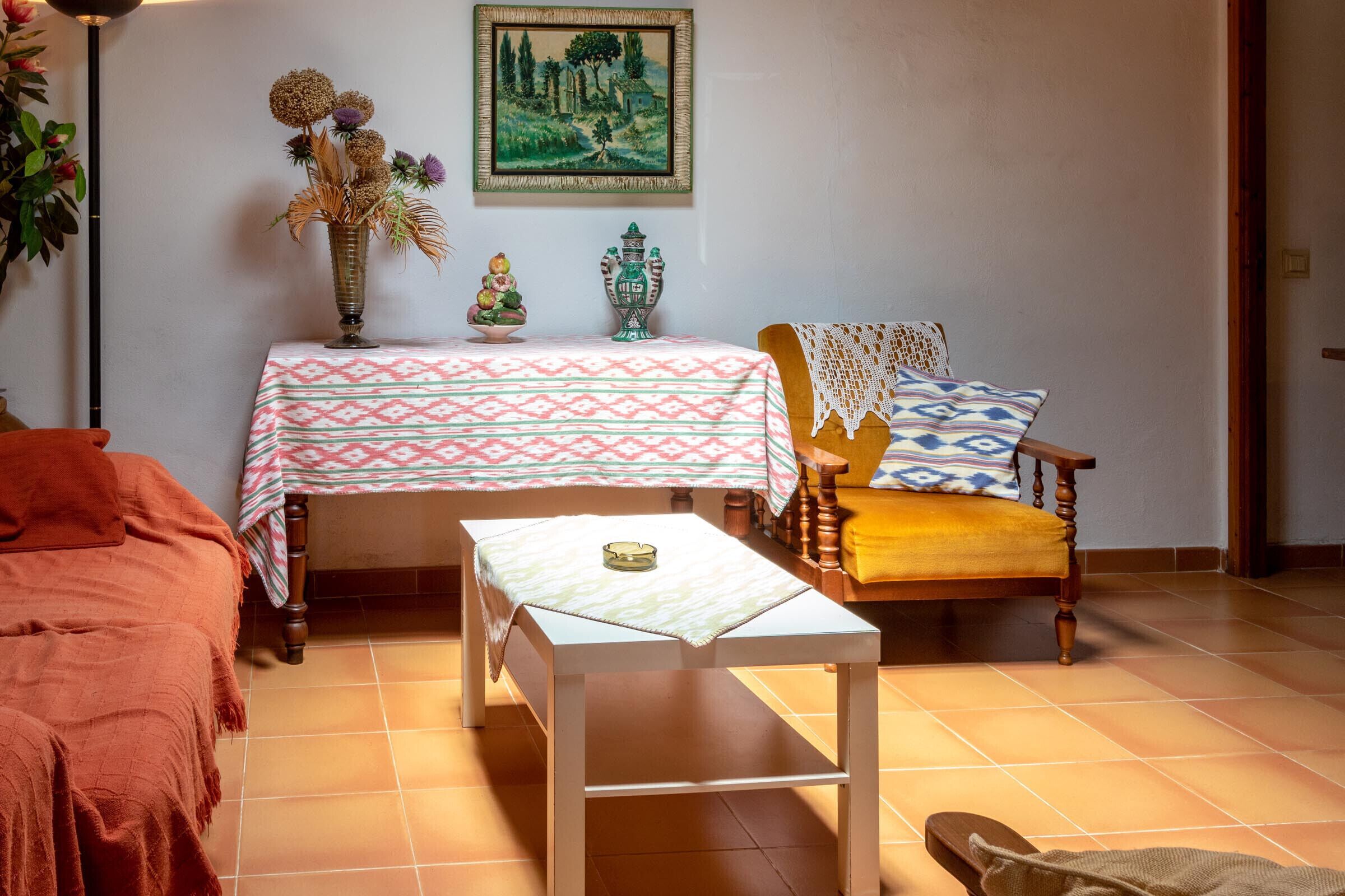 A cozy living room with a patterned table, a wooden chair, and a bed with a red blanket.