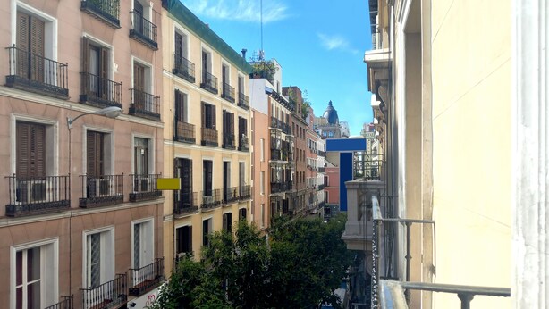 Una hilera de edificios de varios pisos con balcones, un cielo azul despejado y una calle en primer plano.