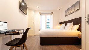 Habitación de hotel con cama, escritorio con una laptop, silla, televisor montado en la pared y ventana con vista a edificios.
