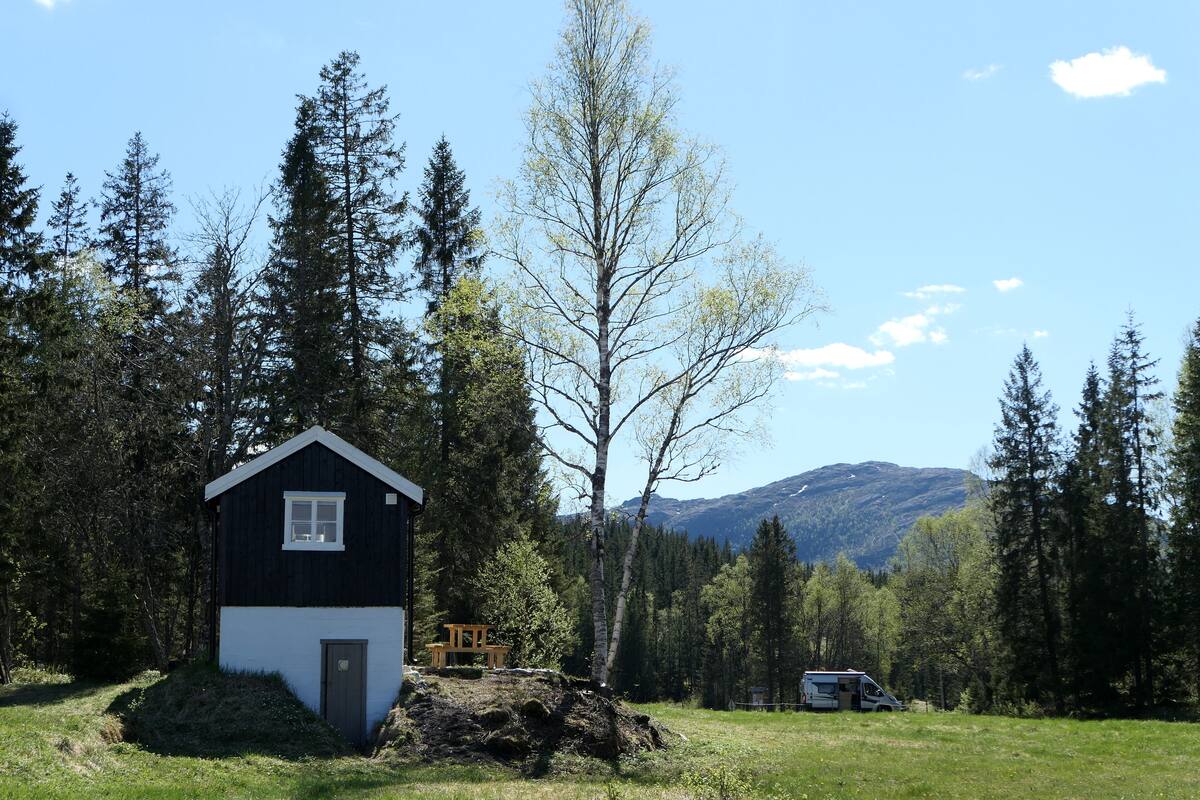 A small cabin with a single window and a door, surrounded by a forest and mountains in the background.