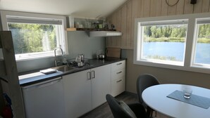 A compact kitchen with a sink, stove, and cabinets, a dining table with chairs, and large windows offering views of a lake and forest. - Velfjord camping & hytter (Brønnøy)