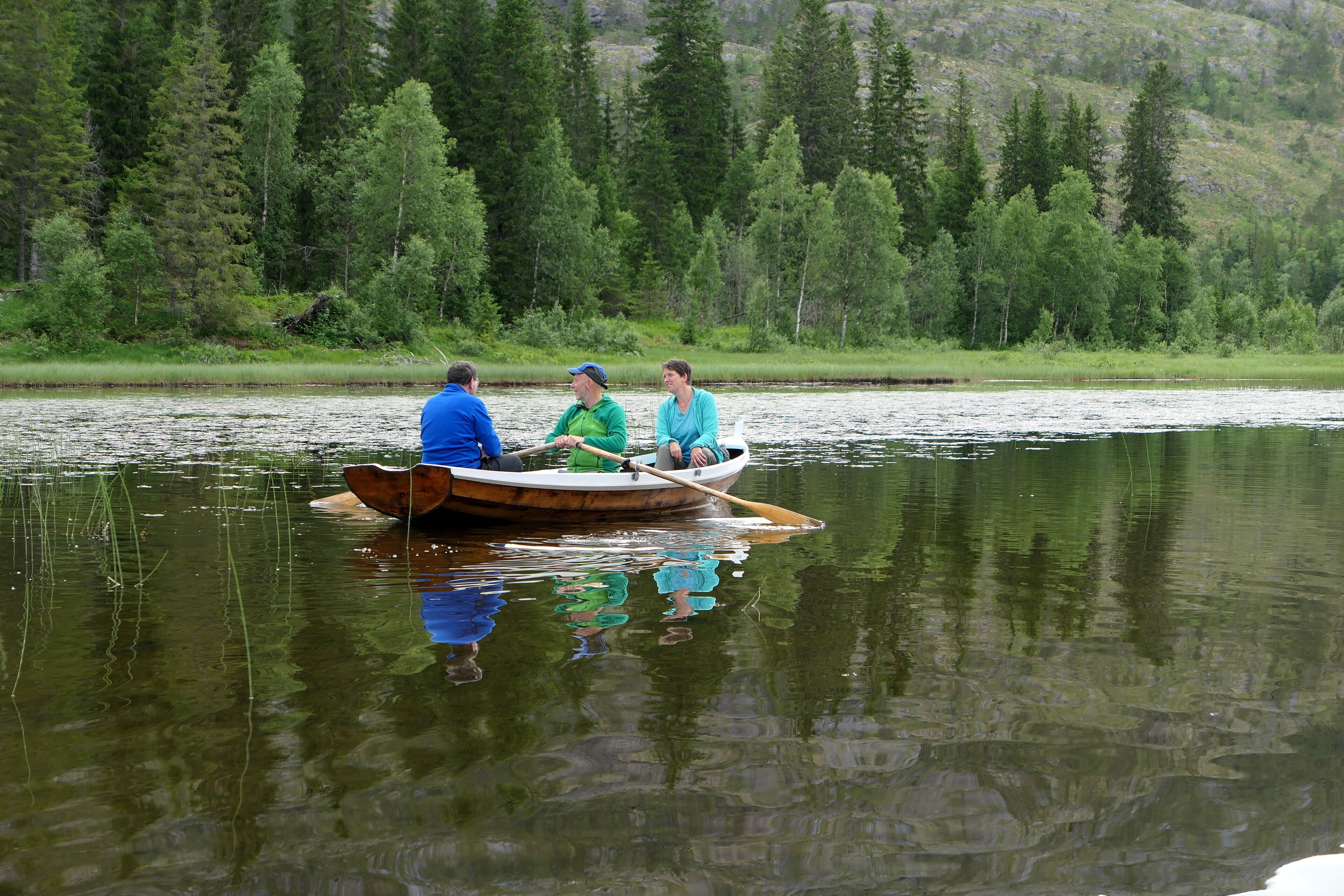 A small wooden boat with three people on a calm lake surrounded by dense forest.