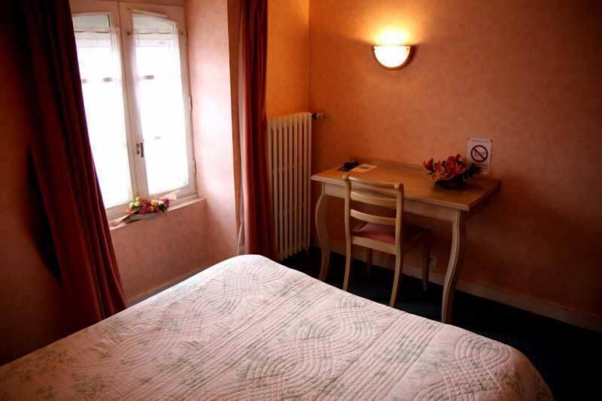 A small bedroom with a bed, a desk, and a chair. There is a window with curtains, a radiator, and a wall-mounted light.