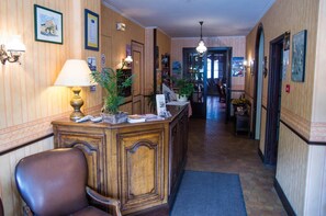 A hotel lobby with a reception desk, a chair, a lamp, and a hallway leading to other rooms. - Hotel de la Gare (Dol-de-Bretagne)