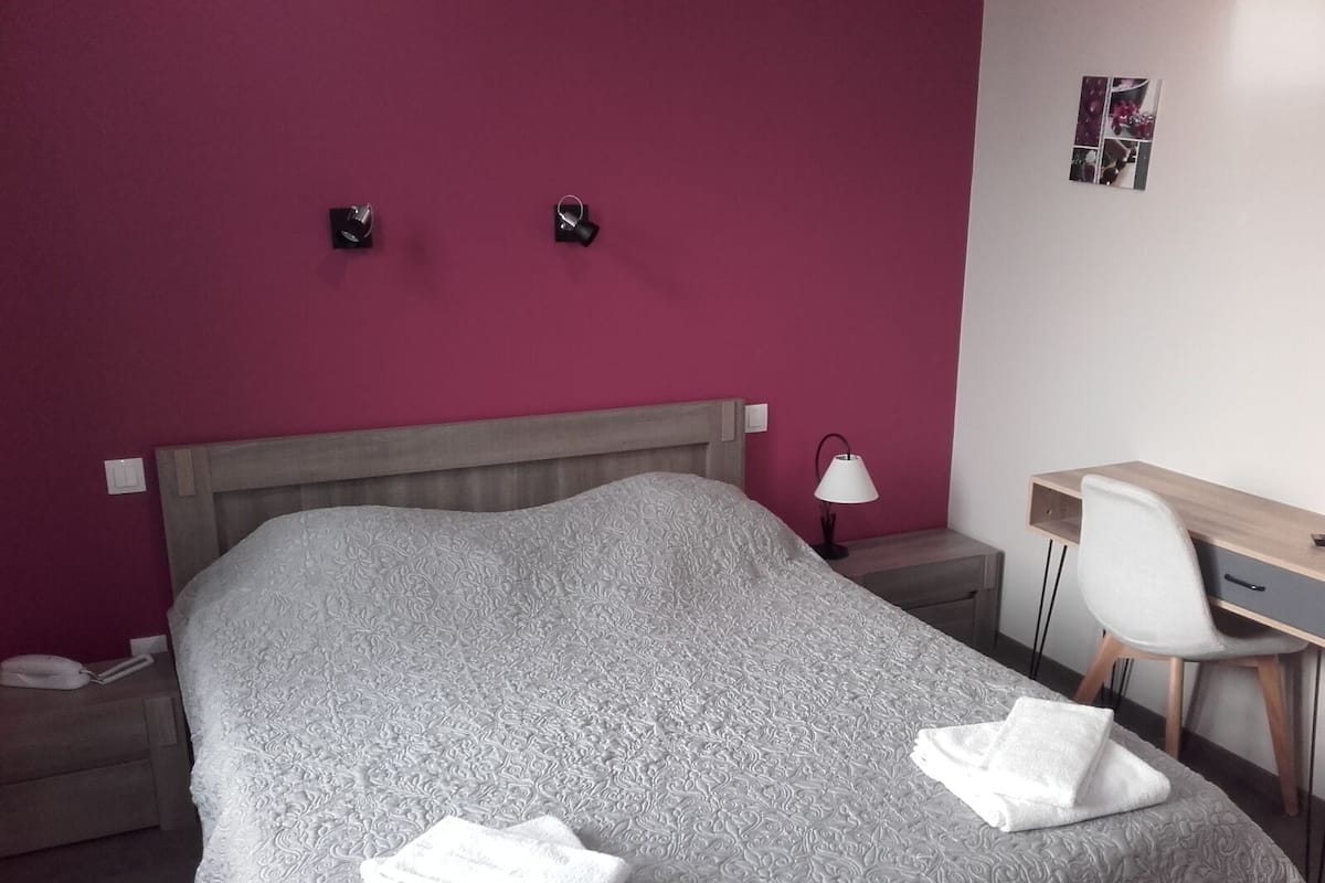 A hotel room with a large bed, a desk, and a chair. There are two wall-mounted lamps and a small table with a lamp.