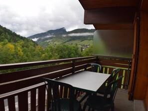 A balcony with a table and chairs, overlooking a valley with houses and mountains.