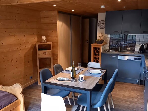 A modern kitchen with a built-in oven, a dining area with a wooden table and blue chairs, and a cozy corner with a wooden cabinet and a heart-shaped drawer.
