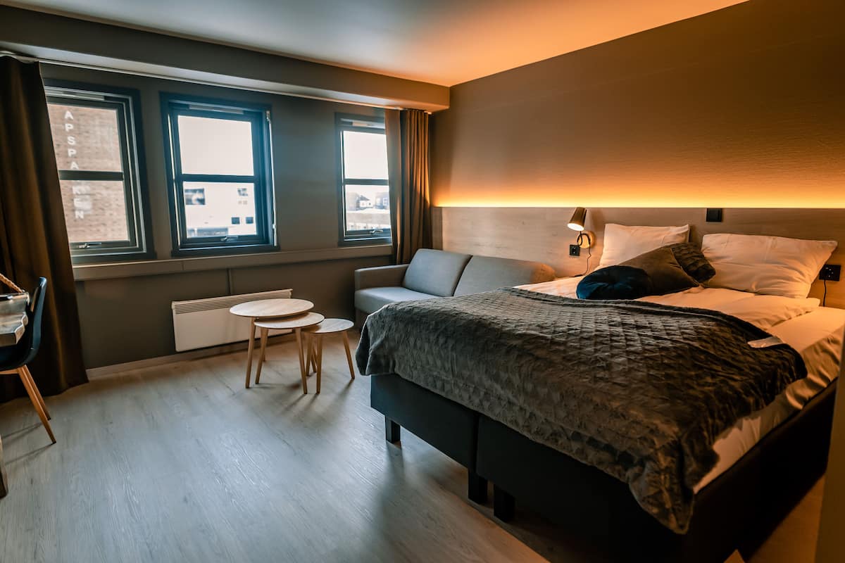 Una habitación de hotel moderna con una cama grande, una mesita redonda pequeña y un sofá. Hay dos ventanas que ofrecen vistas a la ciudad.