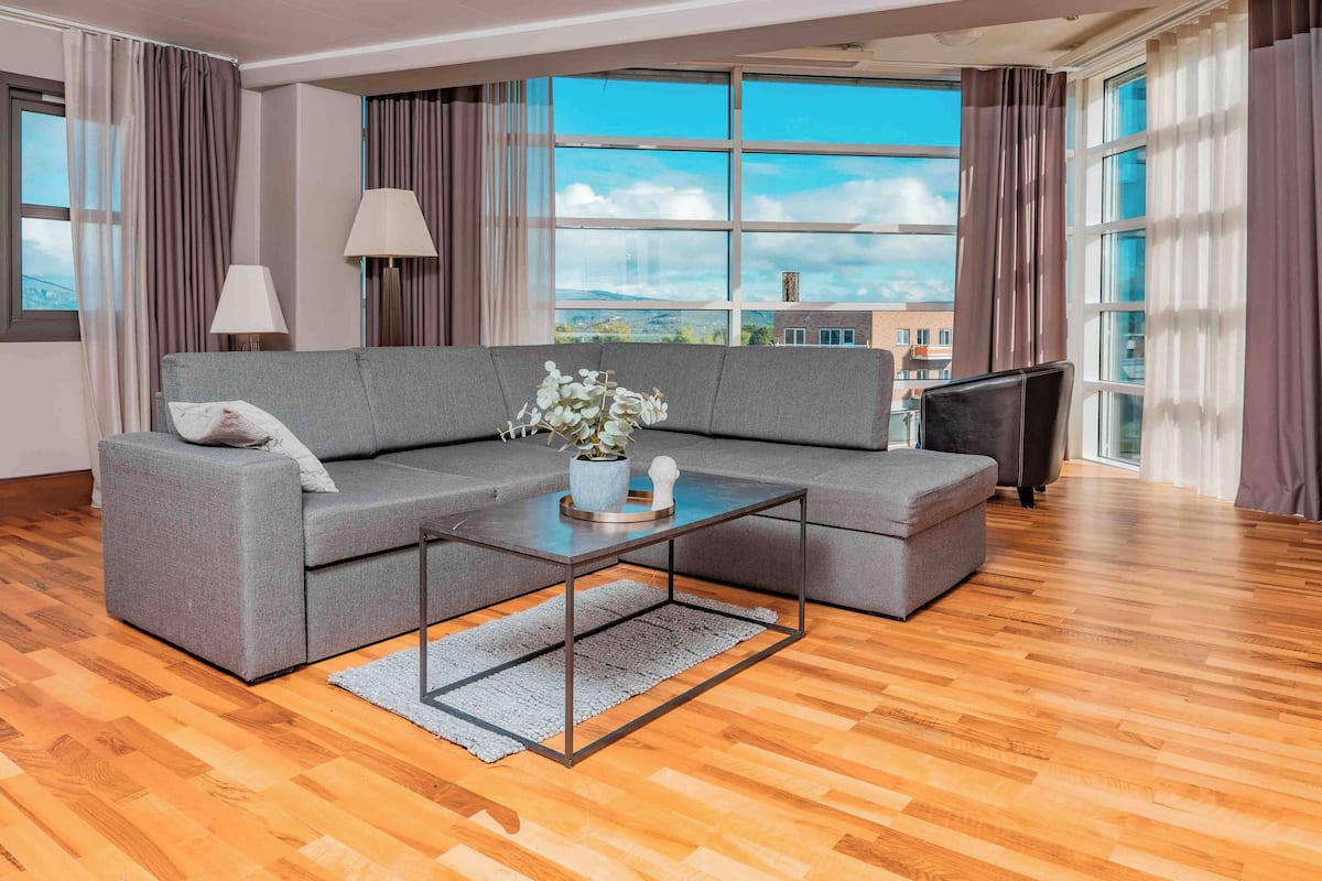 Una sala de estar moderna con un sofá seccional gris, una mesa de centro y un televisor. Ventanas amplias ofrecen vistas del paisaje urbano.