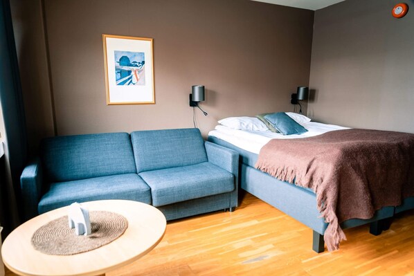 Una habitación de hotel moderna con un sofá, una cama con manta, una mesita redonda con camino de mesa y dos lámparas de pared.