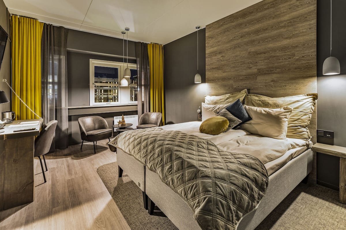 Habitación de hotel moderna con una cama grande, un escritorio y una silla. Cuenta con una ventana con cortinas, una mesita de noche y una lámpara.