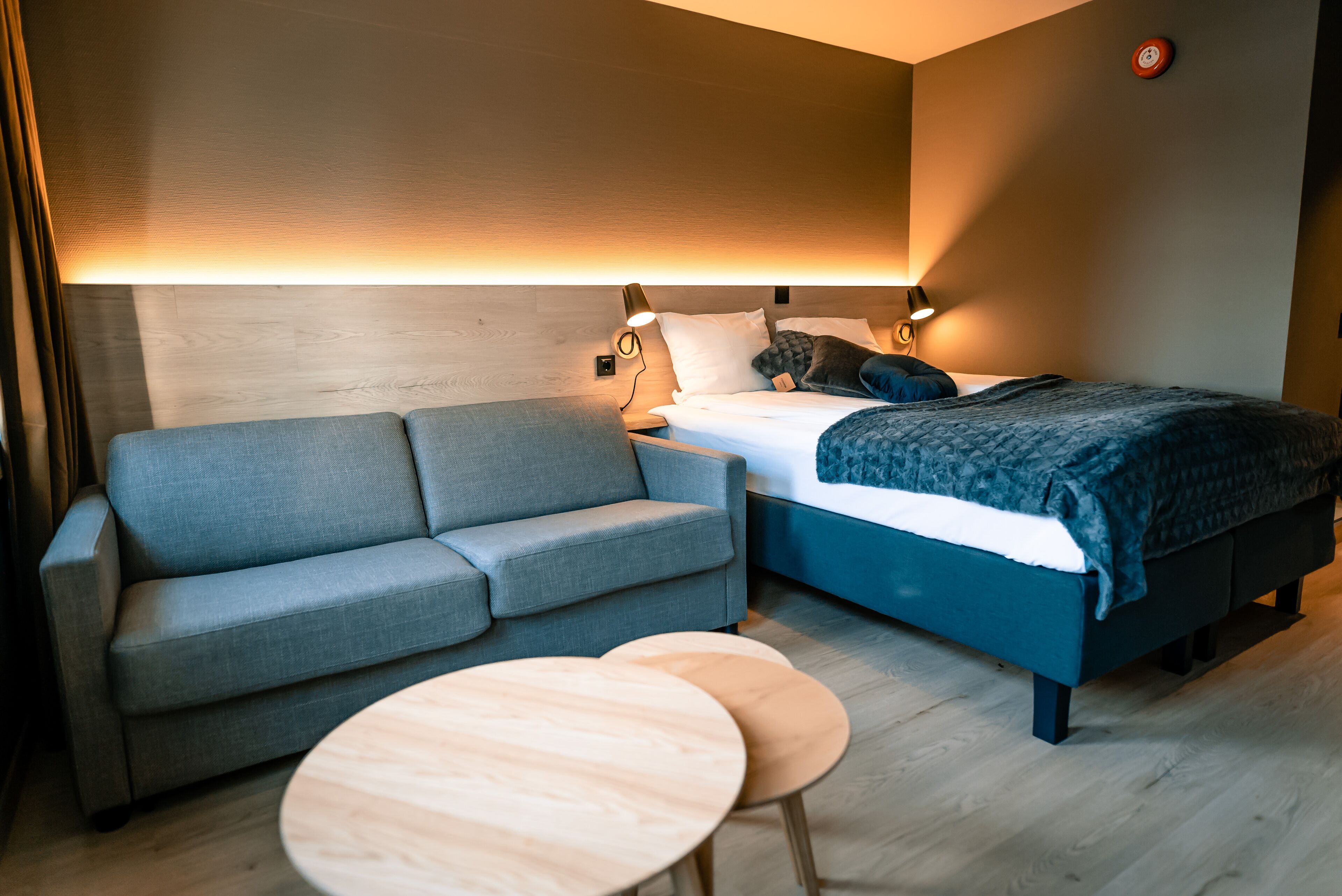 Une chambre d’hôtel moderne avec un canapé gris, un lit doté d’une tête de lit bleue, deux tables de chevet et une lampe fixée au mur.