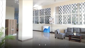 Un hall d’entrée spacieux avec un plafond haut, une grande fenêtre agrémentée de stores à motifs, un comptoir de réception et un espace détente avec des canapés.
