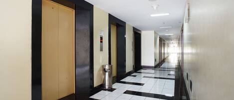 A long, narrow hallway with a checkered floor pattern, a trash can, and a series of doors on one side.