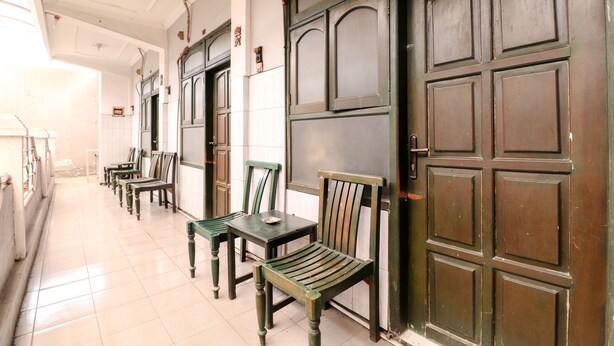 A corridor with wooden benches and tables, a large wooden door, and a tiled floor. - Hotel Gajah Mada (Surabaya)