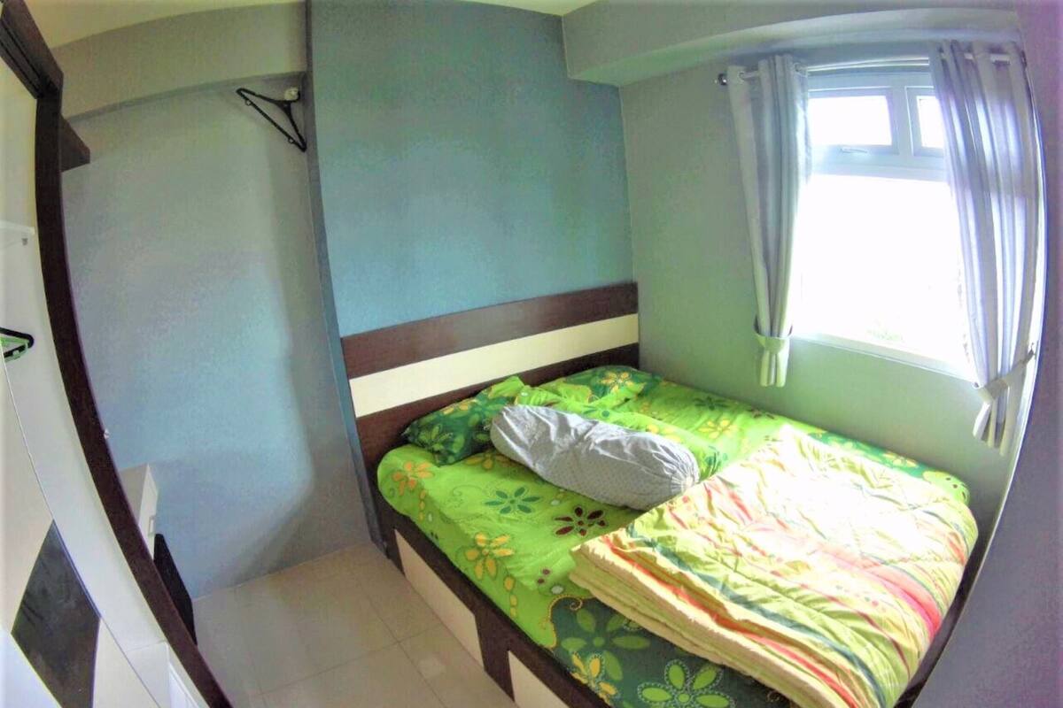 A small, single-bed room with a wooden headboard, a window with curtains, and a wall-mounted shelf.