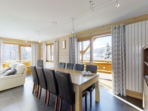 A modern dining area with a wooden table and chairs, large windows with curtains, and a view of snow-covered trees.