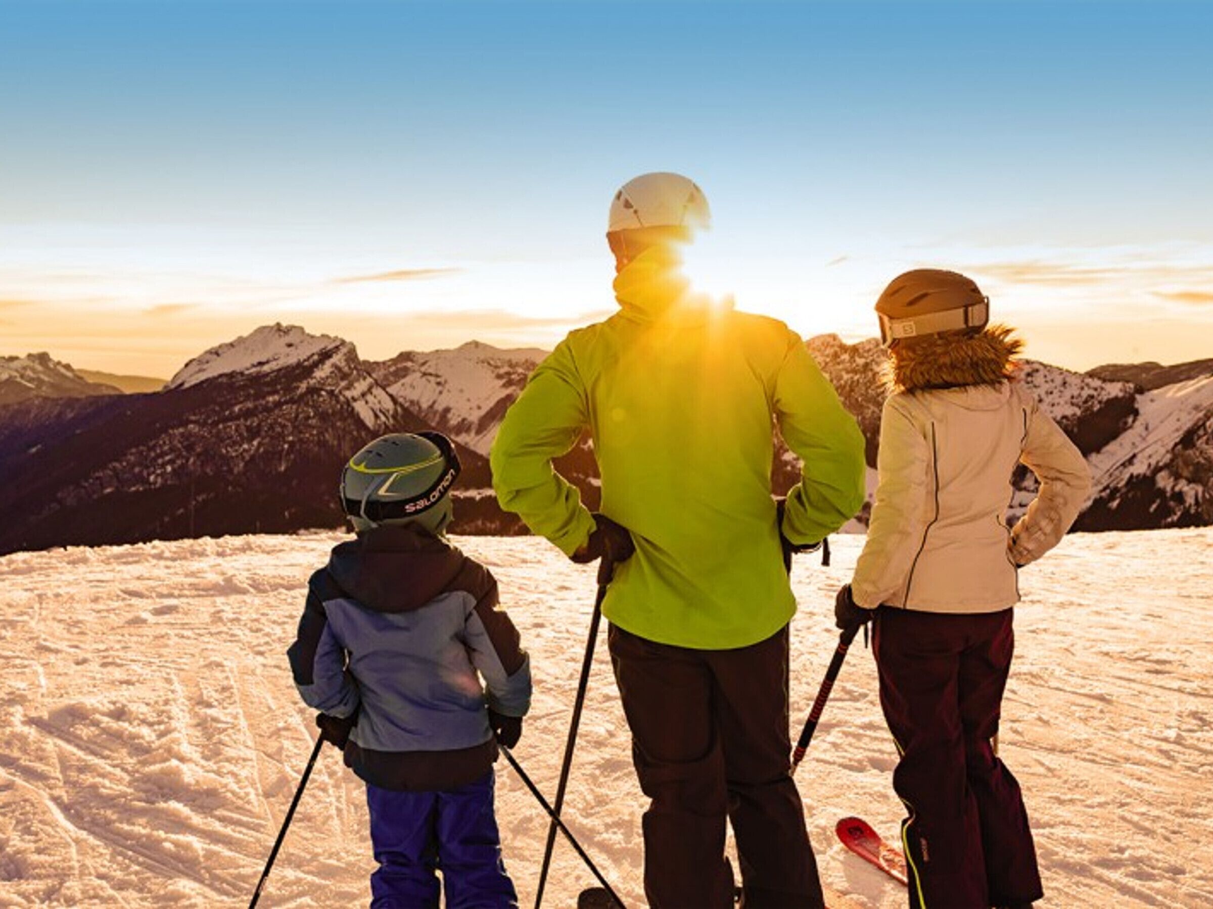 Três pessoas em um monte coberto de neve, com bastões de esqui, apreciando a vista de uma paisagem montanhosa ao pôr do sol.
