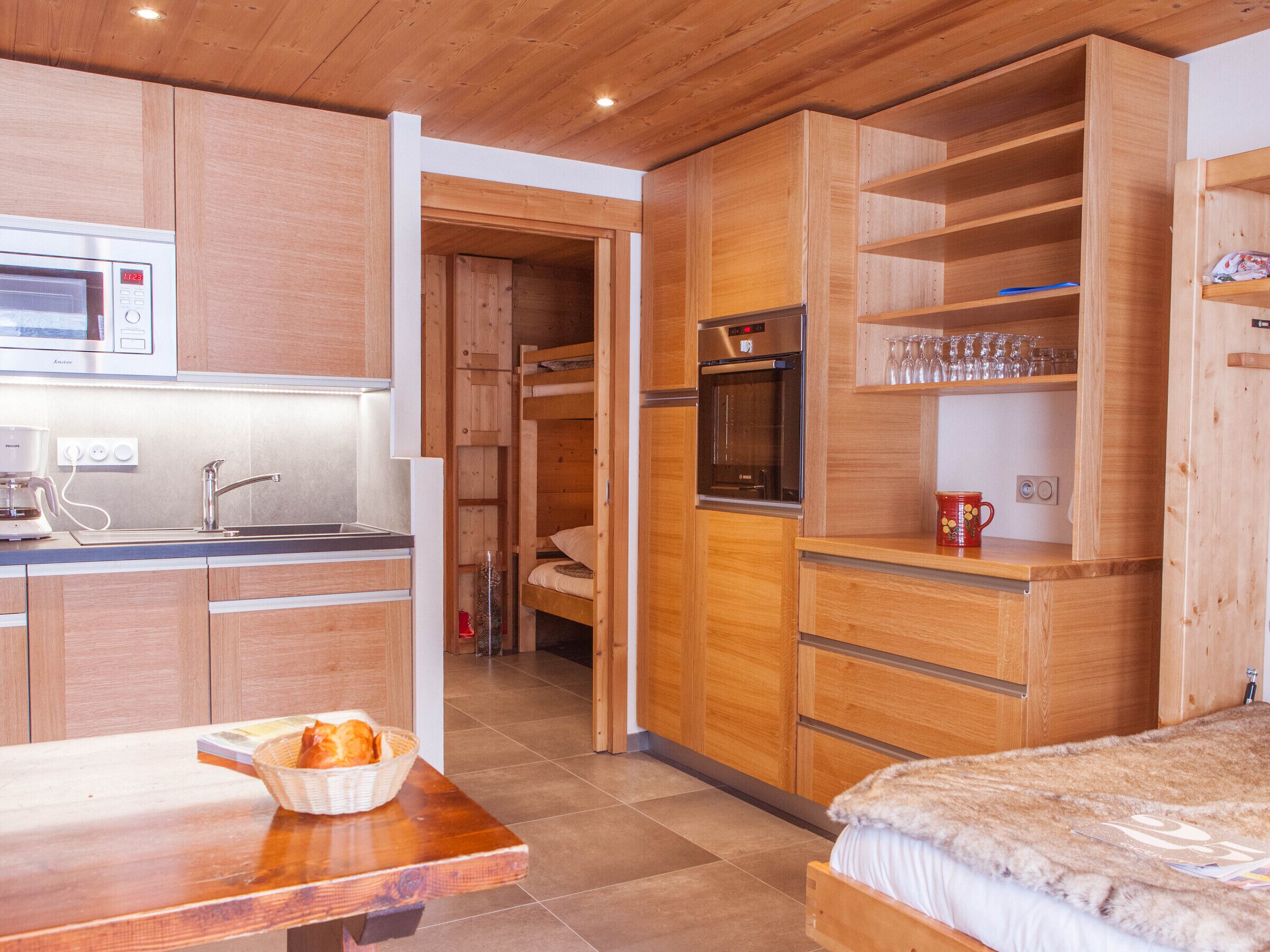 Eine kleine Küche mit Mikrowelle, Kaffeemaschine und Spüle. Ein Holztisch mit einem Korb Brot. Ein Stockbett mit einer flauschigen Decke ist zu sehen.