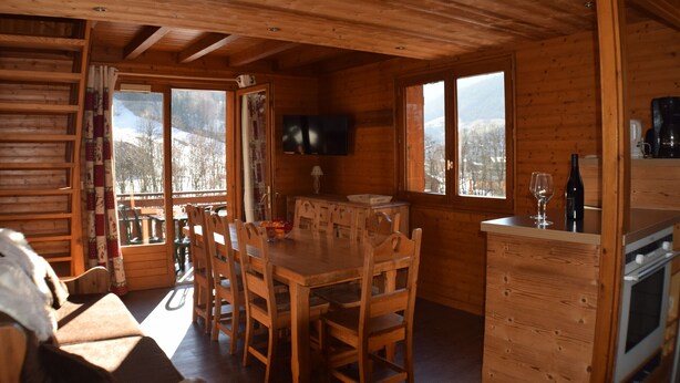 Interior de una cabaña de madera con comedor, cocina y ventanas amplias con vistas a la montaña.