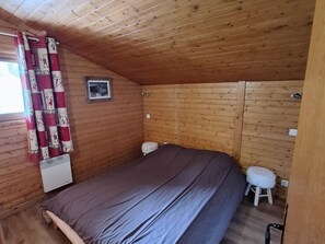 A small, cozy room with wooden walls and a sloped ceiling. It features a single bed with a gray mattress, a small stool, and a radiator.
