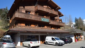Un edificio de madera de varios pisos con balcones, un cartel que indica que es un conjunto de apartamentos y varios coches aparcados delante.