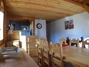 A wooden dining area with a table and chairs, a kitchen in the background, a clock on the wall, and a poster on the wall.