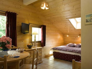 A wooden cabin interior with a bed, a dining table, a lamp, and a TV mounted on the wall.