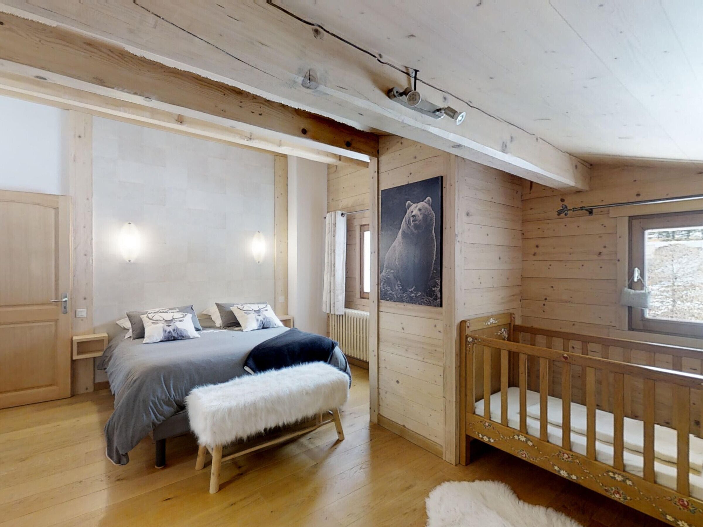Interior de una cabaña de madera con una cama, una cuna, un banco, un radiador y un cuadro de un oso.