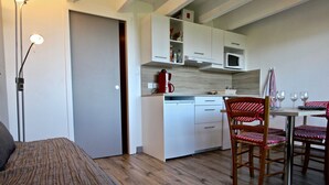 Cozinha compacta com armários brancos, micro-ondas e chaleira vermelha. Possui área de refeições com mesa e cadeiras, e uma cama com colcha listrada.