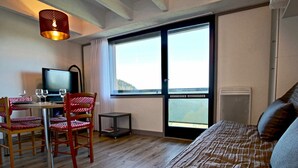 Quarto de hotel com cama, escrivaninha com cadeiras, televisão e uma grande janela.