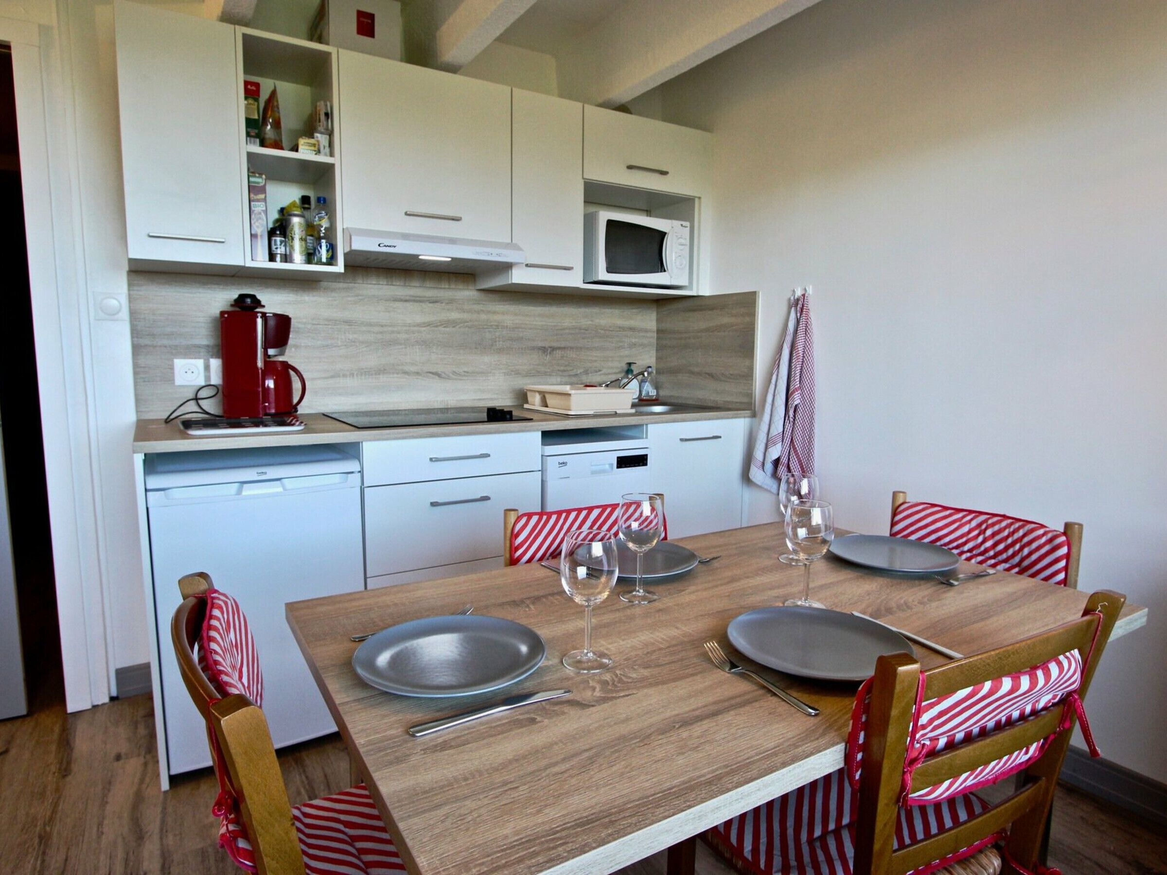 A compact kitchen with white cabinets, a microwave, and a coffee maker. There is a dining table set with plates, wine glasses, and cutlery.
