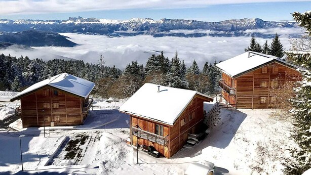 Duas cabanas de madeira com telhados cobertos de neve, com montanhas nevadas e um vale ao fundo.