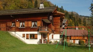 A wooden chalet with a balcony, a staircase leading to the entrance, and a view of the surrounding forest and mountains.