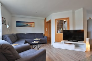 Ein Wohnzimmer mit grauem Sofa, einem kleinen Tisch mit Buch und Tasse, einem Flachbildfernseher auf einem weißen Schrank und einem Bild an der Wand.