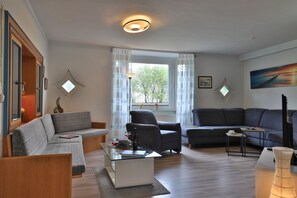 Un salon comprenant un canapé, un fauteuil et une table basse. Il y a une fenêtre avec des rideaux, un tableau au mur et une lampe.