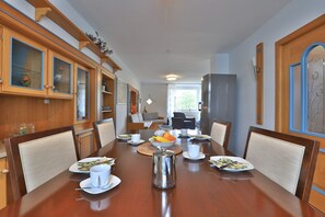 Un coin repas avec une table en bois dressée pour quatre personnes, entourée de chaises. On trouve, à gauche, des armoires avec des portes en verre, à droite, un réfrigérateur, et une fenêtre donnant sur un paysage verdoyant.
