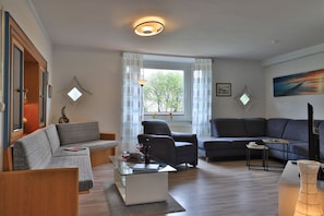 Ein Wohnzimmer mit einer Couch, einem Sessel und einem Couchtisch. Ein Fenster mit Vorhängen, ein Gemälde an der Wand und eine Lampe sind ebenfalls vorhanden.