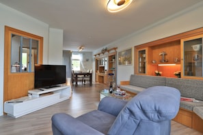 Ein modernes Wohnzimmer mit einem Flachbildfernseher, einem grauen Sofa, Holzschränken und einem Essbereich im Hintergrund.