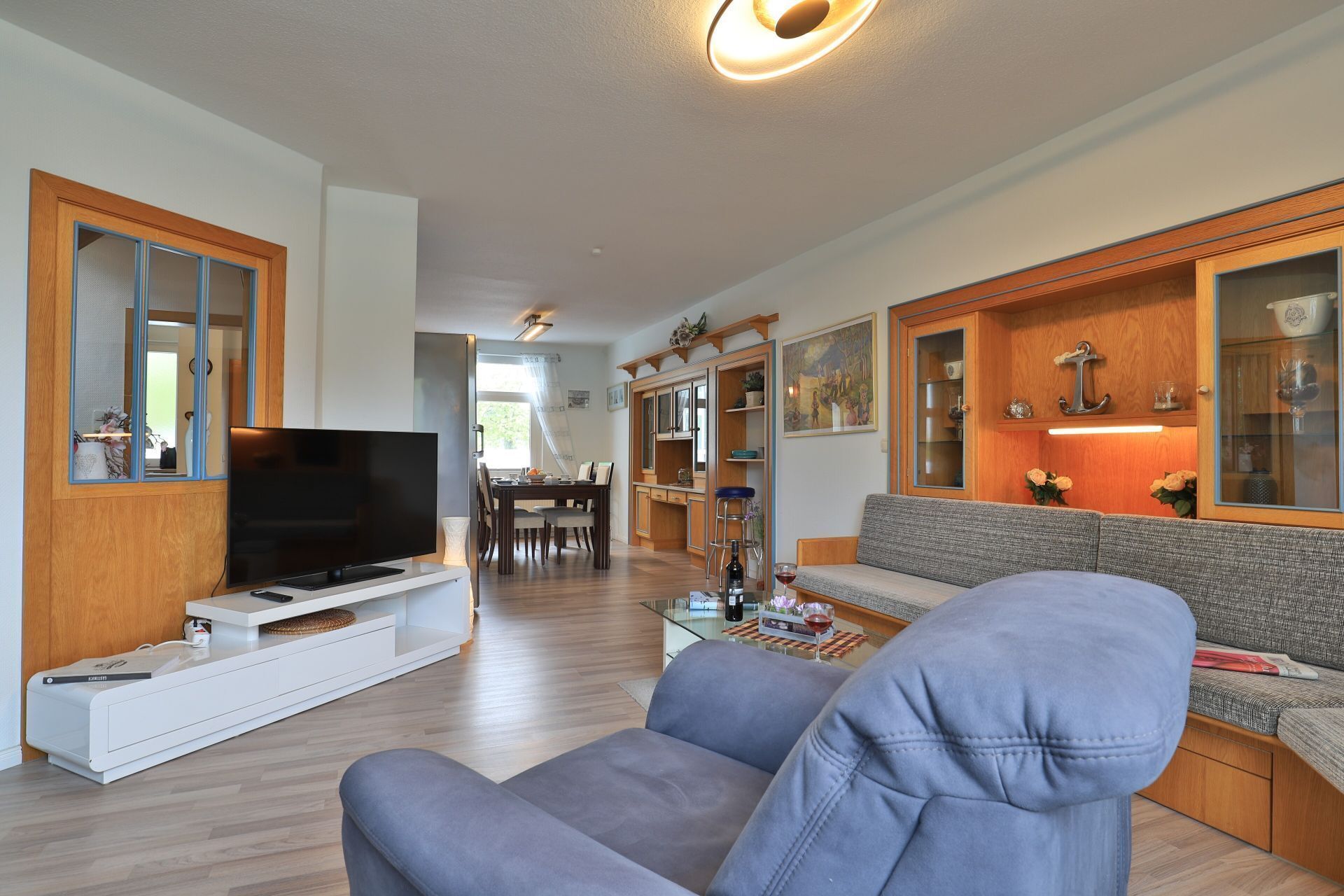 A modern living room with a flat-screen TV, a grey sofa, wooden cabinets, and a dining area in the background.