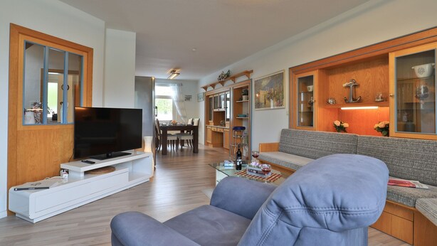 Un salon moderne avec un téléviseur à écran plat, un canapé gris, des meubles en bois et un coin repas à l’arrière-plan.