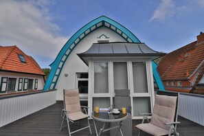 Un edificio moderno con un techo triangular distintivo, un balcón con dos sillas y una mesa, y vistas a casas vecinas.