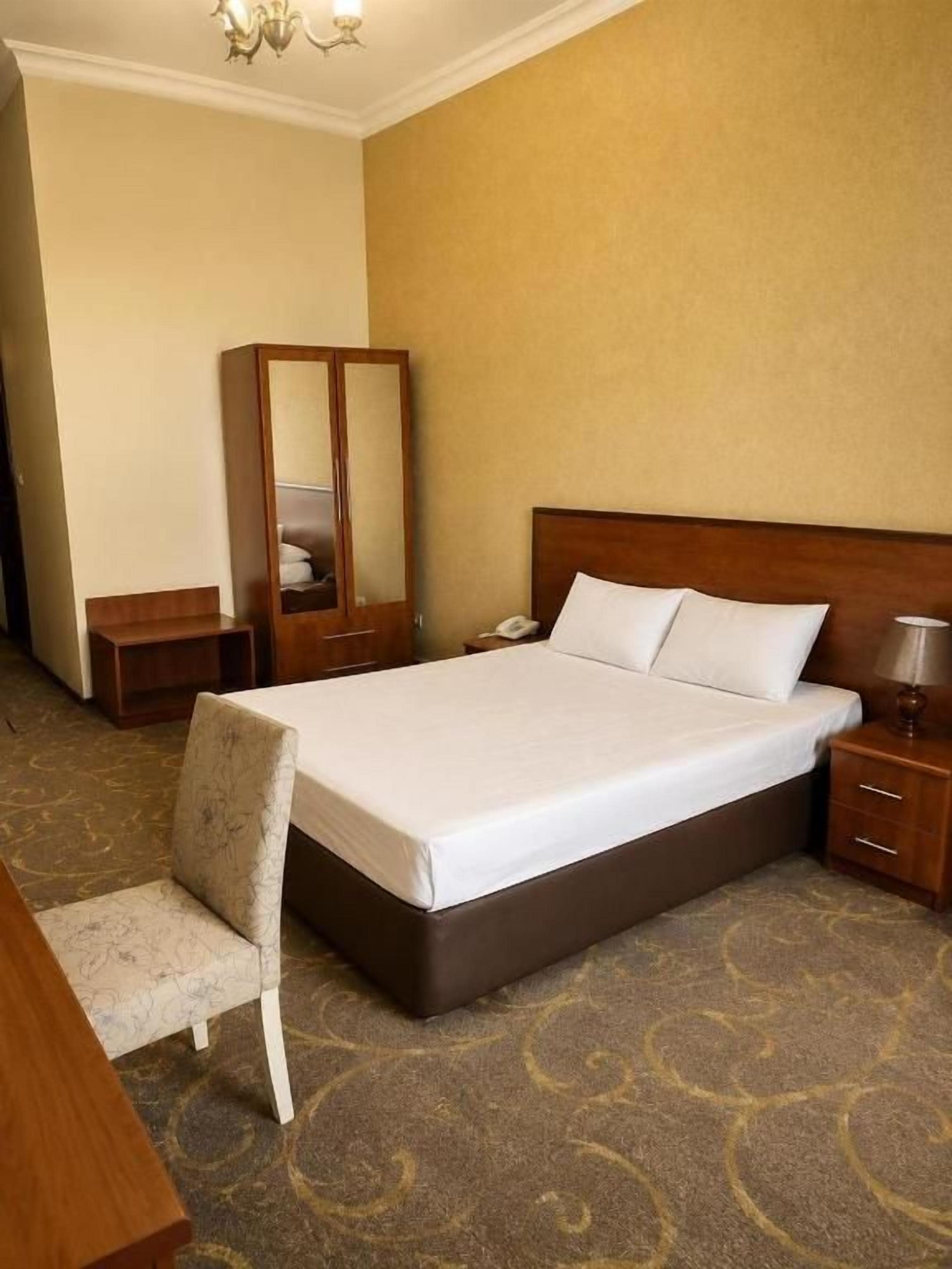 A hotel room with a large bed, a bedside table, a chair, a wardrobe, and a carpeted floor.