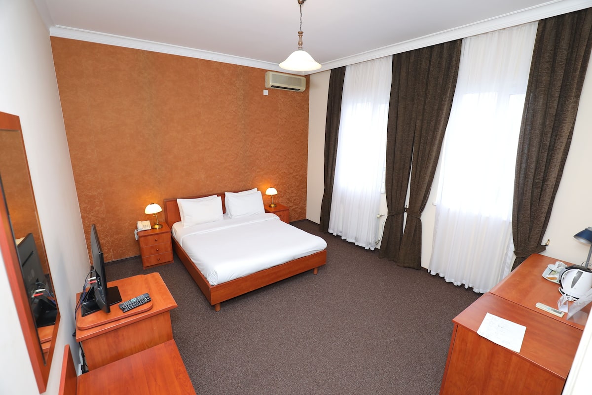 A hotel room with a large bed, a desk with a chair, a TV, and a desk lamp.