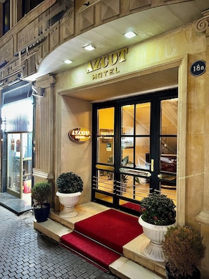 L’extérieur de l’hôtel Azcot, avec son entrée imposante, son tapis rouge et ses plantes en pot.