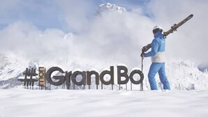 A person standing in the snow with skis, in front of a sign that reads "GrandBo".